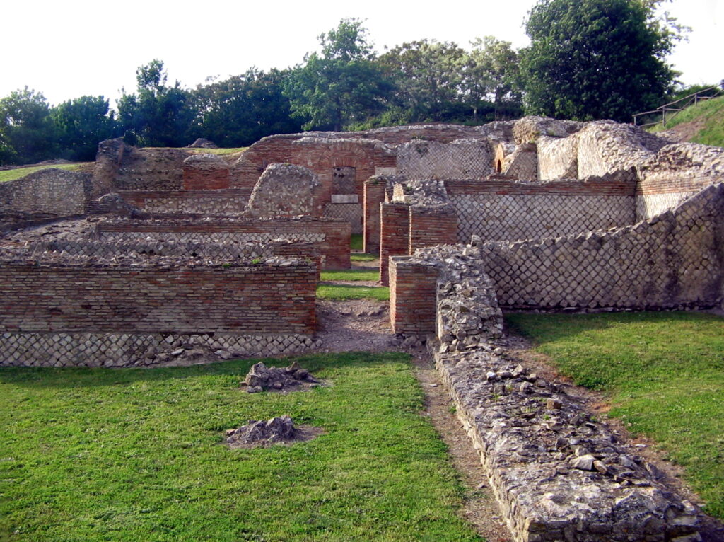 Ruins of Aeclanum, a Roman town in Irpinia district, now Avellino, Campania. The ruin stands in a green park with trees in the background. A diamond patterning is visible on most of the walls, alongside thinly layered horizontal reddish bricks.