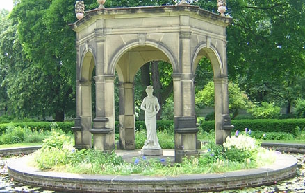 Detail from photo of a statue to Harmonia in the Harmony Society gardens, Old Economy Village (now Ambridge), PA. The statue is photographed from a distance; it is in the centre of an ornate hexagonal stone gazebo surrounded by a ring of wild flowers, on a stand in the middle of a pond. Harmonia is depicted standing, head slightly bowed, holding what appears to be a lyre.