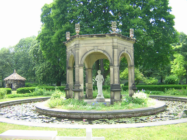 A statue to Harmonia in the Harmony Society gardens, Old Economy Village (now Ambridge), PA. The statue is photographed from a distance; it is in the centre of an ornate hexagonal stone gazebo surrounded by a ring of wild flowers, on a stand in the middle of a pond. Harmonia is depicted standing, head slightly bowed, holding what appears to be a lyre. Trees, hedges and a rustic hut are shown in the background.
